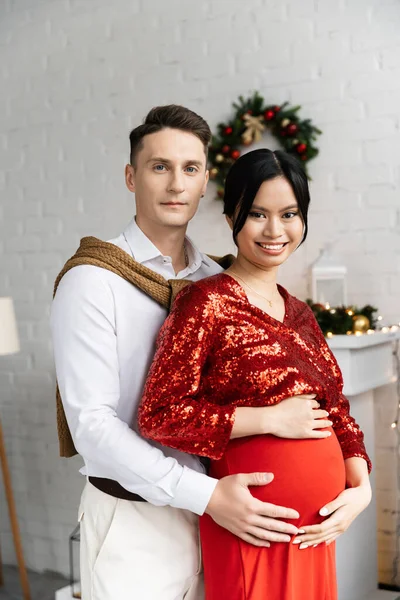 Incinta ed elegante asiatico donna sorridente a macchina fotografica vicino marito abbracciando il suo ventre — Foto stock