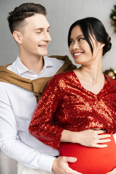Homem feliz e mulher asiática grávida em roupas festivas sorrindo um para o outro — Fotografia de Stock