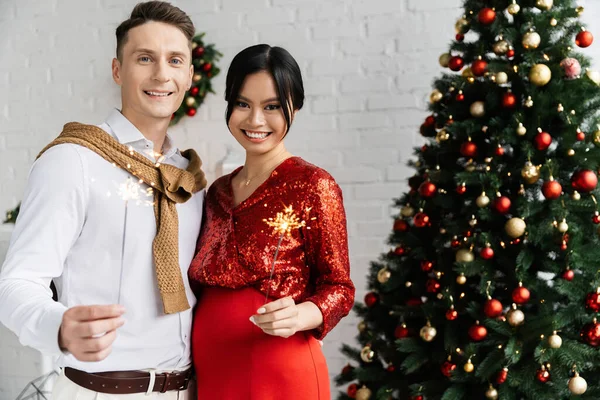 Homem feliz com grávida asiático esposa segurando brilhante sparklers perto decorado árvore de Natal em casa — Fotografia de Stock