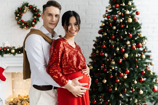 Schwangere und glückliche Asiatin mit Ehemann, der ihren Bauch in der Nähe des geschmückten Weihnachtsbaums umarmt — Stockfoto
