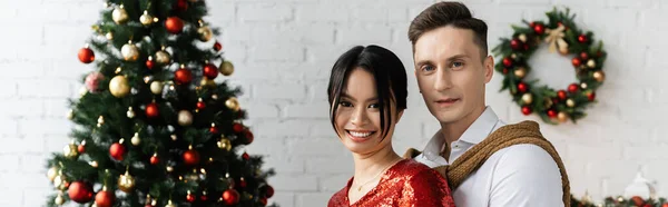 Happy and elegant interracial couple looking at camera near blurred christmas tree, banner — Stock Photo
