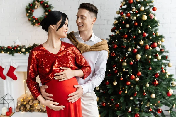 Cheerful man and pregnant asian woman in elegant clothes smiling at each other in living room with Christmas decor — Stock Photo