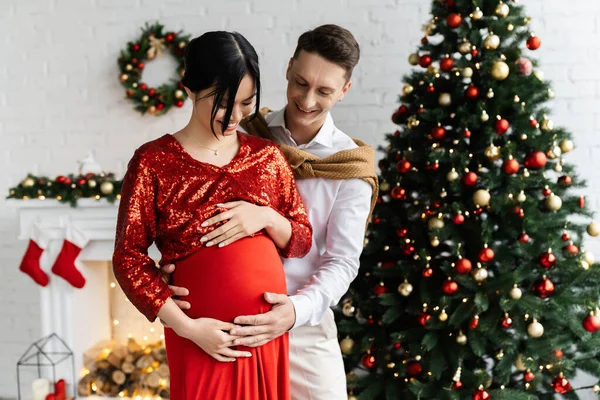 Uomo sorridente che abbraccia pancia di incinta moglie asiatica in soggiorno con arredamento natalizio — Foto stock