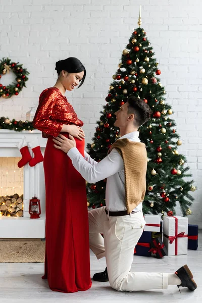 Vista laterale dell'uomo in piedi sul ginocchio e abbracciando incinta ed elegante moglie asiatica vicino all'albero di Natale — Foto stock