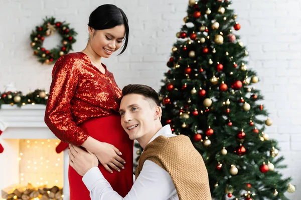 Счастливый мужчина опираясь на животик беременной и элегантной азиатской женщины возле елки — стоковое фото
