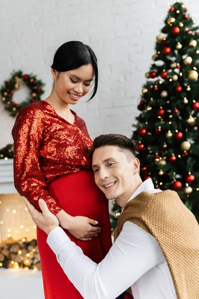Homem alegre inclinando-se na barriga de grávida asiática esposa perto borrada árvore de Natal em casa — Fotografia de Stock