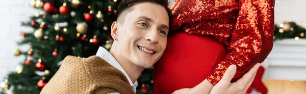 Усміхнений чоловік обіймає животик вагітної дружини біля різдвяної прикраси на розмитому фоні, банер — стокове фото