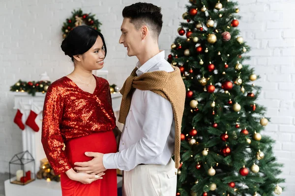 Junger Mann und schwangere Asiatin in eleganter Kleidung schauen sich nahe geschmücktem Weihnachtsbaum an — Stockfoto
