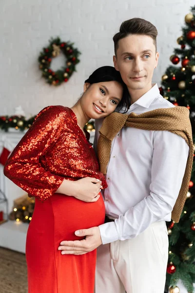 Pregnant asian woman in festive clothes smiling near husband and Christmas decor on blurred background — Stock Photo