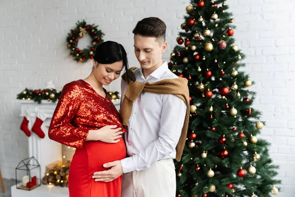 Young man touching belly of pregnant asian wife in festive clothes in living room with Christmas decor — Stock Photo