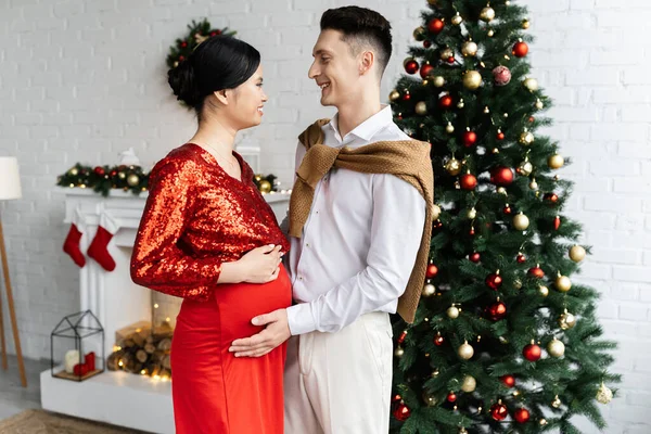 Side view of pregnant and elegant asian woman with husband smiling at each other near Christmas tree in living room — Stock Photo