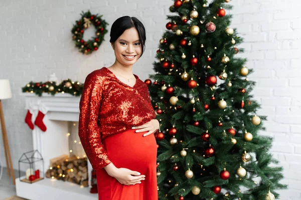 Alegre y embarazada asiático mujer en rojo elegante ropa mirando cámara cerca decorado árbol de Navidad - foto de stock