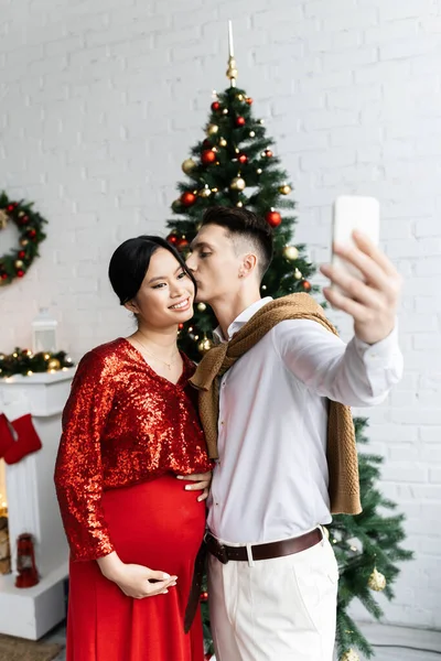 Jovem homem beijos elegante e grávida ásia mulher enquanto tomando selfie perto borrado Natal árvore — Fotografia de Stock
