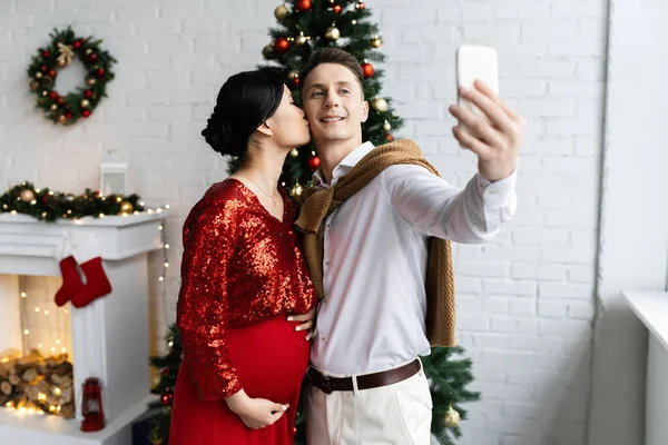 Вагітна і елегантна азіатська жінка цілує чоловіка, приймаючи селфі на мобільний телефон в декорованій вітальні — стокове фото
