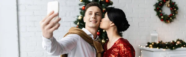 Молода азіатка цілує веселого чоловіка, який бере селфі на смартфон біля розмитого різдвяного декору, банер — стокове фото
