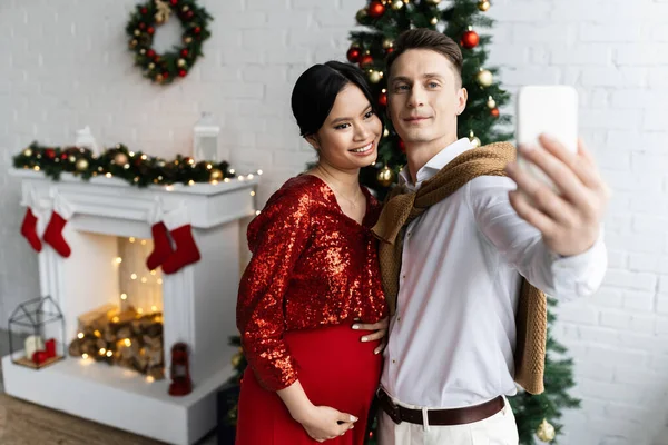 Jovem tomando selfie com feliz e grávida asiático esposa perto de natal árvore e decorado lareira — Fotografia de Stock