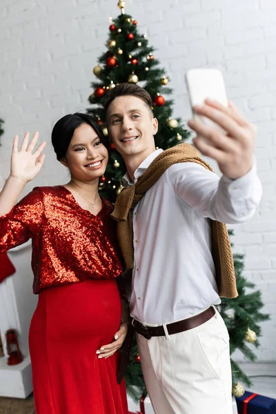 Happy man with mobile phone near pregnant asian wife waving hand during video call on Christmas day — Stock Photo
