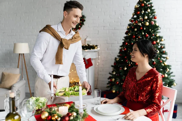 Sorridente uomo che serve pasto vicino incinta moglie asiatica seduta a tavola con romantica cena di Natale — Foto stock