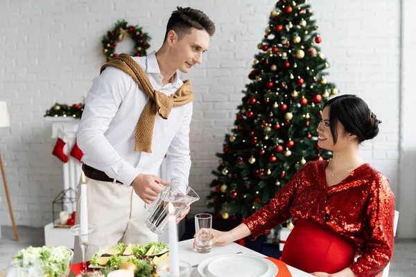 Giovane uomo versando acqua in vetro vicino elegante e incinta asiatico moglie a romantico Natale cena — Foto stock