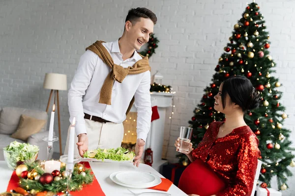 Uomo sorridente che serve pasto vicino moglie asiatica incinta con un bicchiere d'acqua durante la cena di Natale — Foto stock