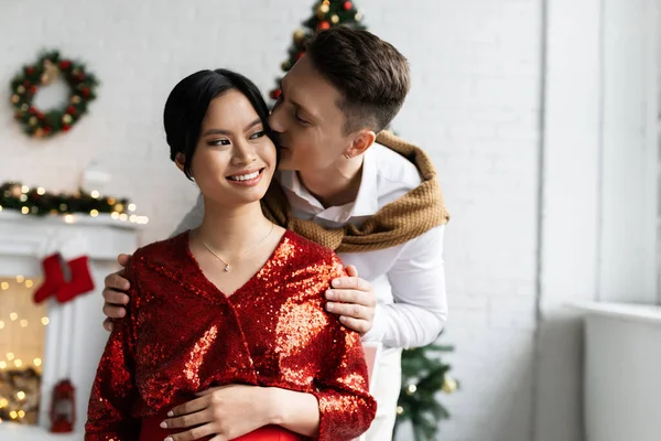 Молодой человек целует беременную азиатку в красной и блестящей блузке возле рождественского декора на размытом фоне — стоковое фото