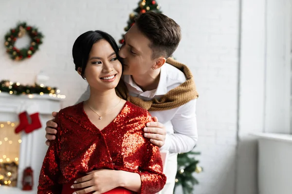 Young man kissing pregnant asian woman in red and shiny blouse near Christmas decor on blurred background — Stock Photo