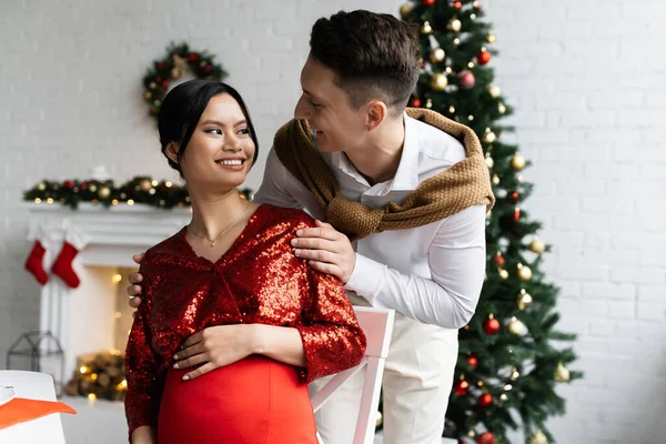 Jovem homem olhando grávida asiático esposa sentado no sala de estar com Natal decoração — Fotografia de Stock