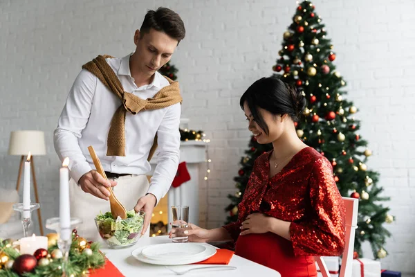 Pregnant asian woman smiling near man with fresh vegetable salad during romantic Christmas supper at home — Stock Photo
