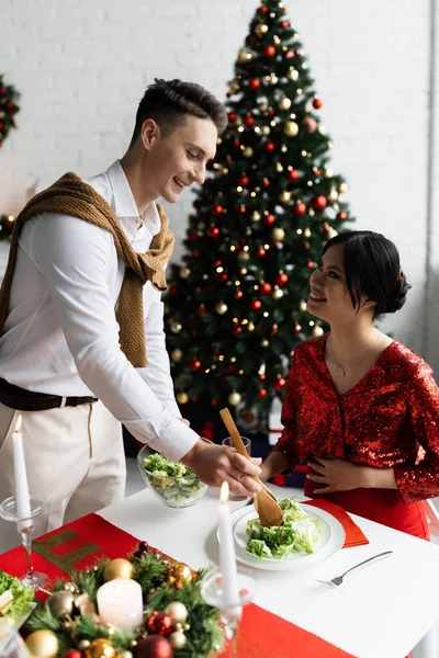 Беременная азиатка смотрит на счастливого мужа, подающего овощной салат возле горящих свечей на романтическом рождественском ужине — стоковое фото