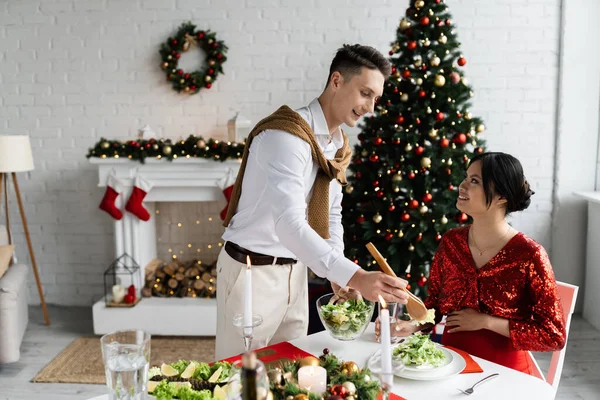 Schwangere Asiatin schaut Ehemann mit Schüssel Gemüsesalat in der Nähe von dekoriertem Kamin und Weihnachtsbaum an — Stockfoto