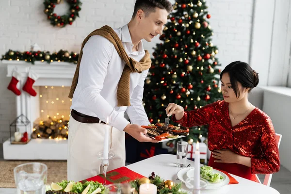 Hombre sosteniendo a la parrilla verduras cerca embarazada y elegante asiático mujer durante romántica cena cerca de árbol de Navidad - foto de stock