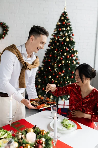 Souriant homme tenant assiette avec des légumes grillés près asiatique femme avec fourchette pendant romantique souper de Noël — Photo de stock