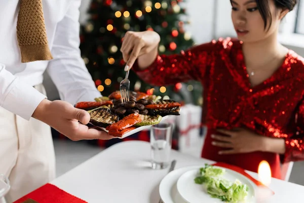 Hombre sosteniendo a la parrilla verduras cerca embarazada asiático esposa con tenedor durante romántico Navidad cena sobre borrosa fondo - foto de stock