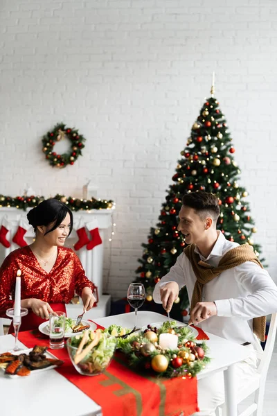 Feliz hombre y su embarazada asiático esposa sonriendo el uno al otro mientras que tener Navidad cena en casa - foto de stock