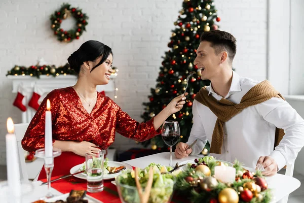 Pregnant and cheerful asian woman feeding husband while having fun at romantic Christmas supper — Stock Photo