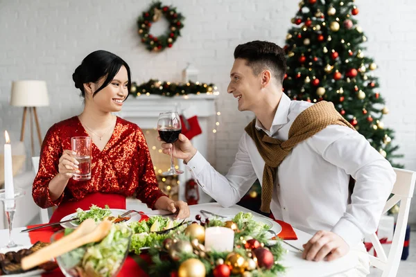 Grávida asiática mulher com marido segurando óculos com bebidas durante romântico Natal ceia em sala de estar — Fotografia de Stock