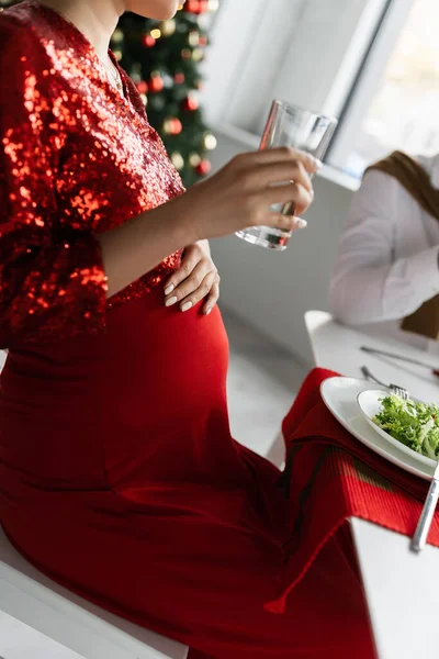 Vista recortada de la mujer embarazada sosteniendo un vaso de agua cerca de la lechuga fresca durante la cena de Navidad con el marido - foto de stock