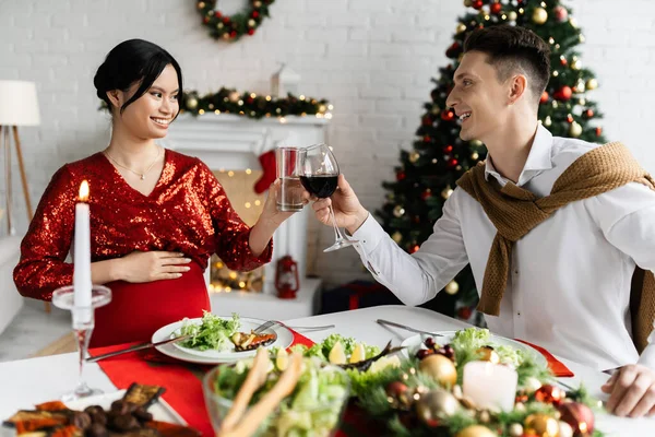 Happy man clinking glasses with pregnant and elegant asian wife near table served with festive Christmas dinner — Stock Photo