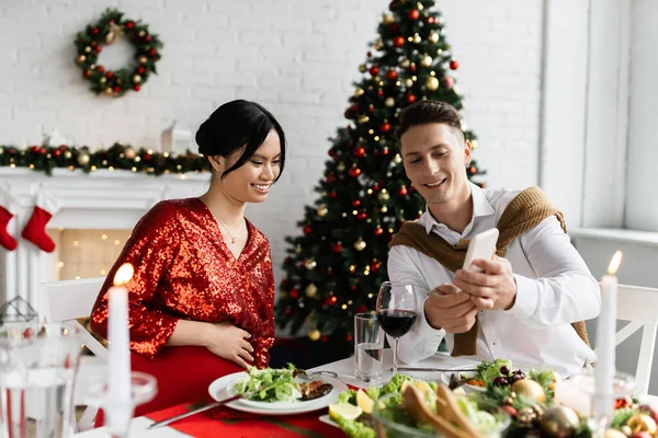 Smiling man showing smartphone to pregnant asian wife during romantic Christmas supper at home — Stock Photo