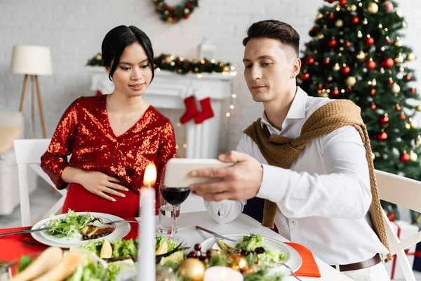 Young man showing smartphone to pregnant asian woman near served supper in living room with Christmas tree — Stock Photo