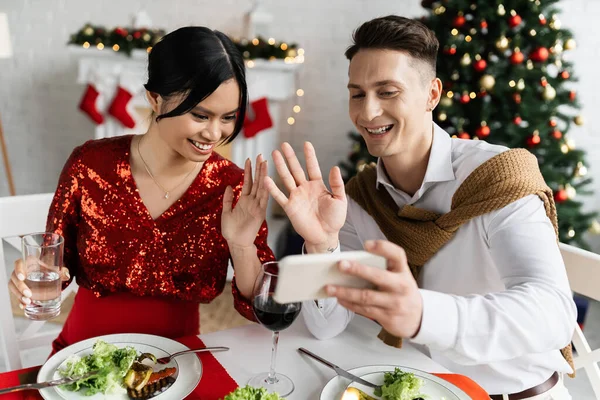 Pregnant asian woman with smiling husband waving hands during video call on smartphone at Christmas celebration — Stock Photo
