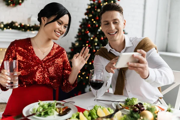 Homme souriant tenant smartphone près enceinte asiatique femme agitant la main lors de l'appel vidéo lors du souper festif — Photo de stock