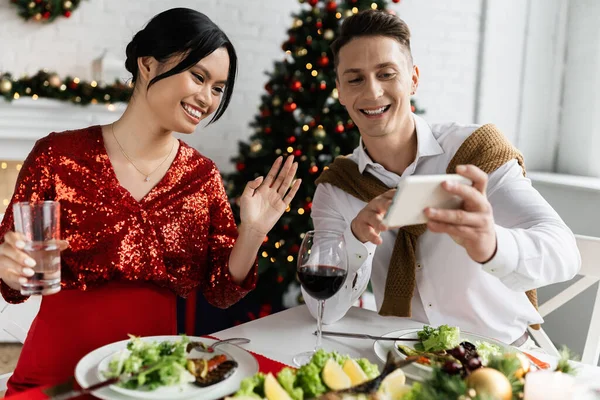 Smiling man holding smartphone near pregnant asian wife waving hand during video call during festive supper — Stock Photo
