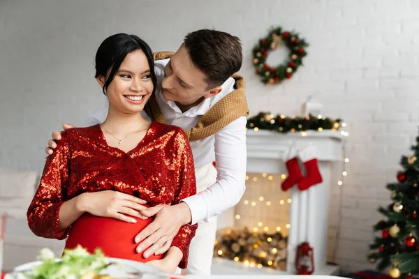 Uomo toccare pancia di incinta asiatico moglie in elegante abiti durante natale celebrazione — Foto stock