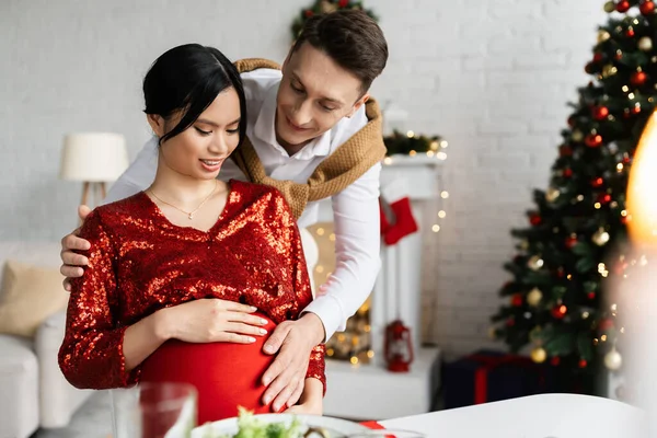 Lächelnder Mann berührt Bauch einer schwangeren Asiatin, während er zu Hause Weihnachten feiert — Stockfoto