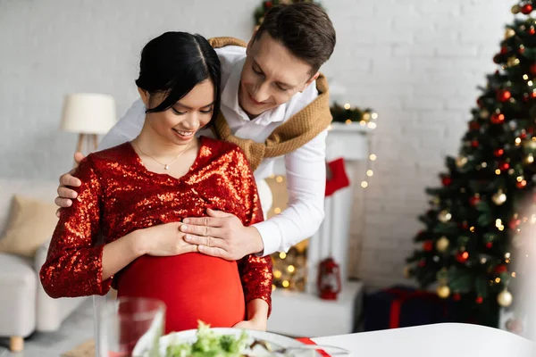 Happy and pregnant asian woman in elegant clothes sitting near husband touching her belly in decorated living room — Stock Photo