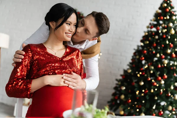 Giovane uomo baci allegro e incinta asiatico moglie vicino offuscata albero di Natale a casa — Foto stock