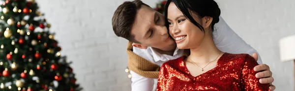 Jovem homem beijar feliz asiático esposa no vermelho e brilhante blusa perto borrada árvore de Natal, banner — Fotografia de Stock
