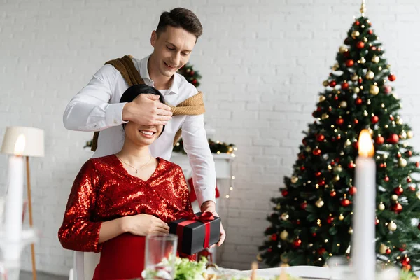 Fröhlicher Mann bedeckt Augen von schwangerer asiatischer Frau, während er Weihnachtsgeschenk hält — Stockfoto