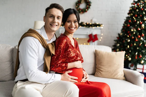 Giovane uomo con incinta asiatico moglie in festa vestito sorridente a macchina fotografica su divano vicino offuscata albero di Natale — Foto stock